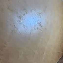 Ściana z dekoracyjną, beżowo-złotą fakturą w mieszkaniu. Widoczny fragment podłogi z paneli i biały sufit z listwą.