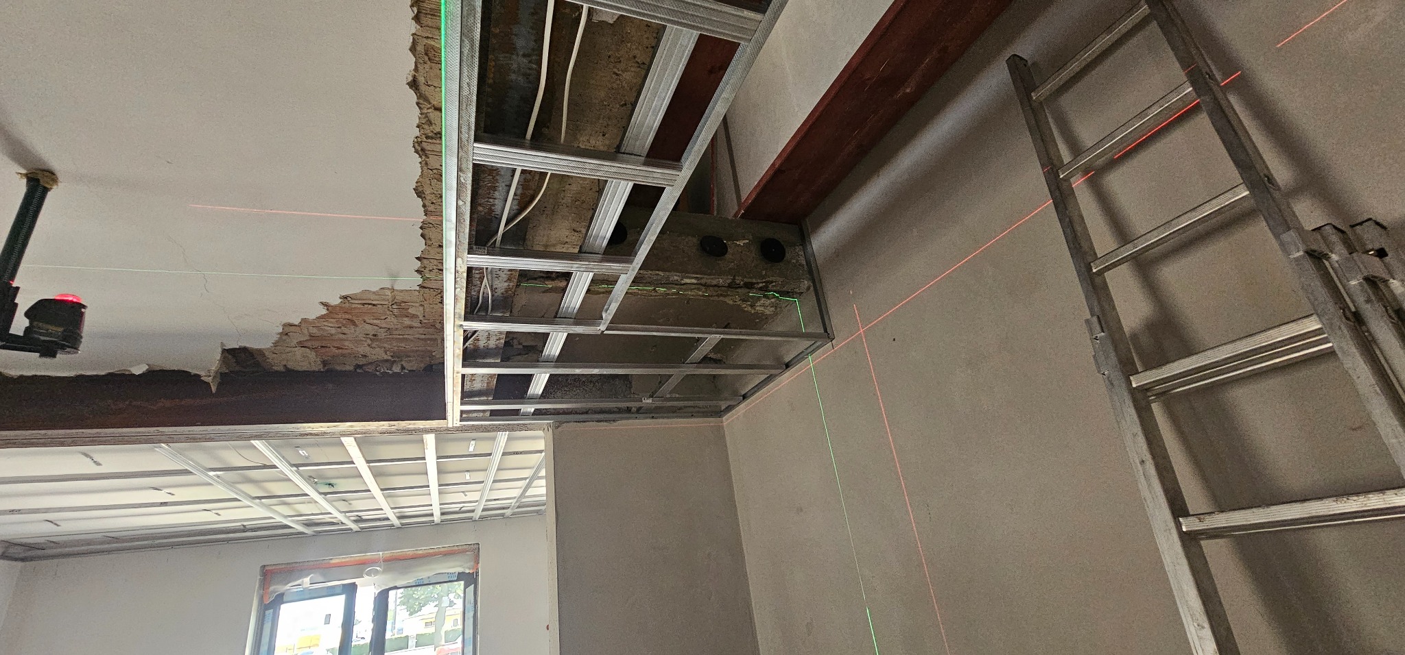 Montaż metalowej konstrukcji sufitu podwieszanego z widocznymi przewodami, poziomica laserowa i drabina oparta o ścianę. W tle okno.