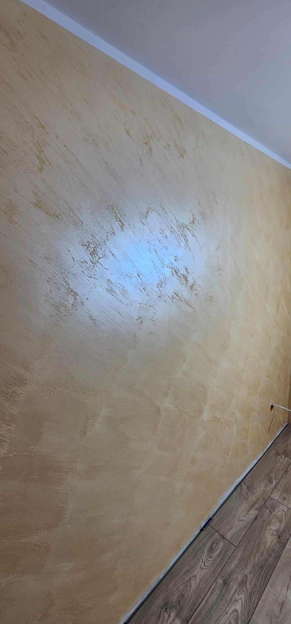 Ściana z dekoracyjną, beżowo-złotą fakturą w mieszkaniu. Widoczny fragment podłogi z paneli i biały sufit z listwą.