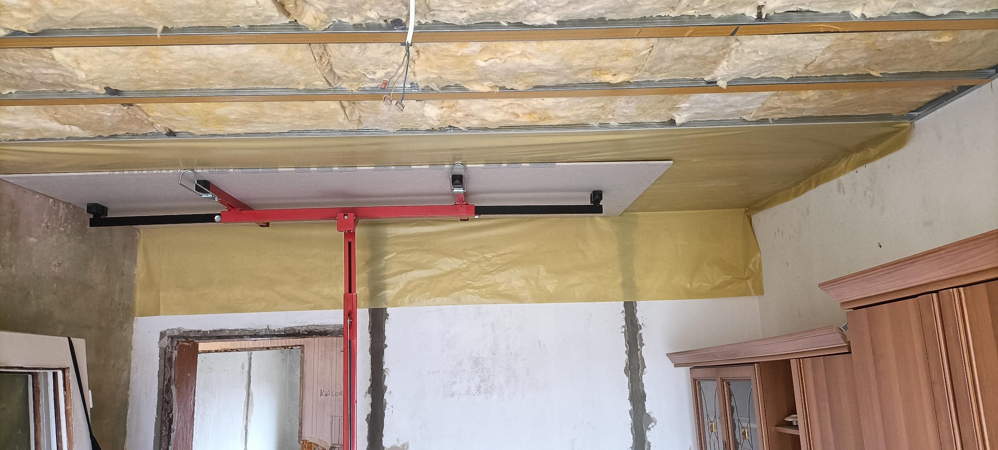 Montaż sufitu podwieszanego z płyty gipsowej za pomocą podnośnika. Widoczne ocieplenie i folia paroizolacyjna. Etap prac wykończeniowych.