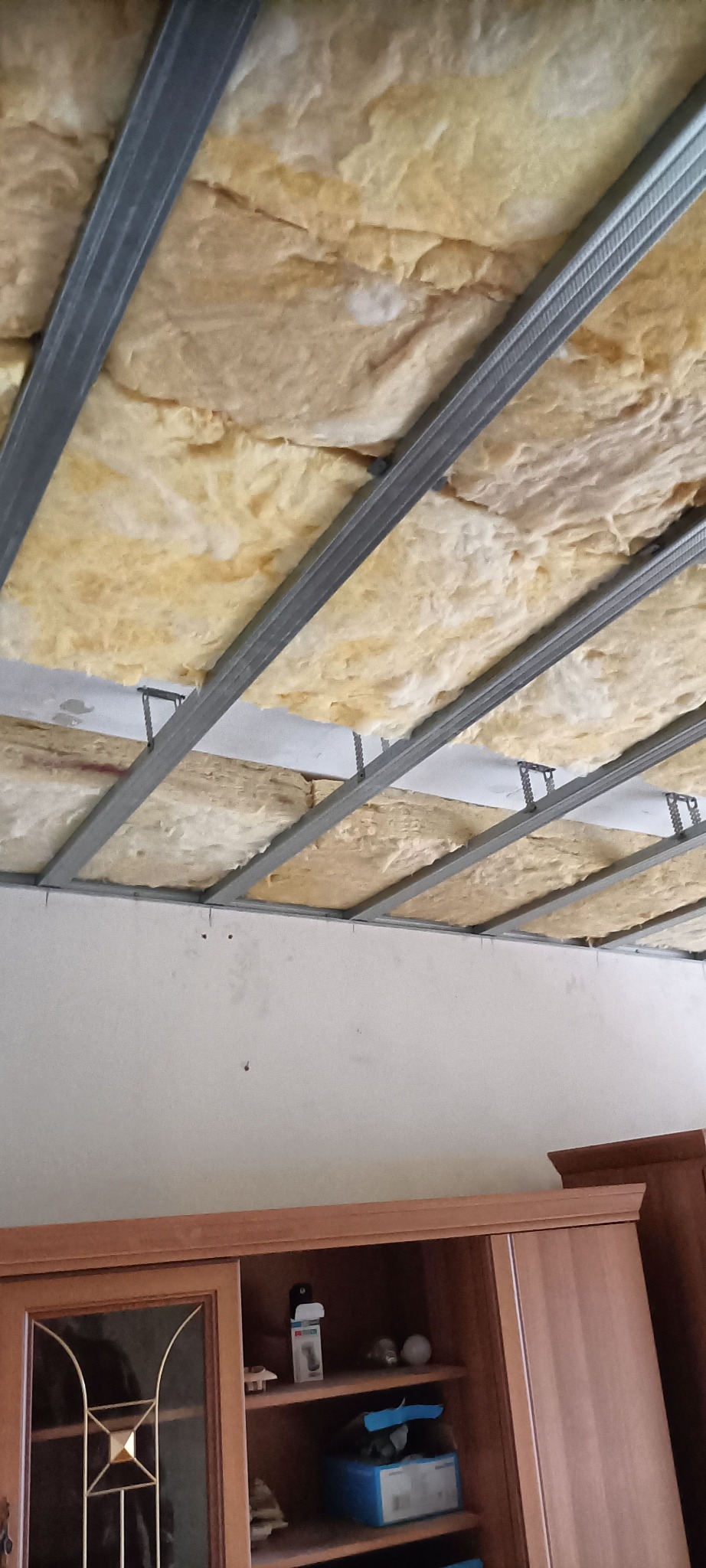Izolacja stropu wełną mineralną z widoczną konstrukcją metalową, przygotowana do montażu sufitu podwieszanego. Widoczny fragment ściany i mebli.