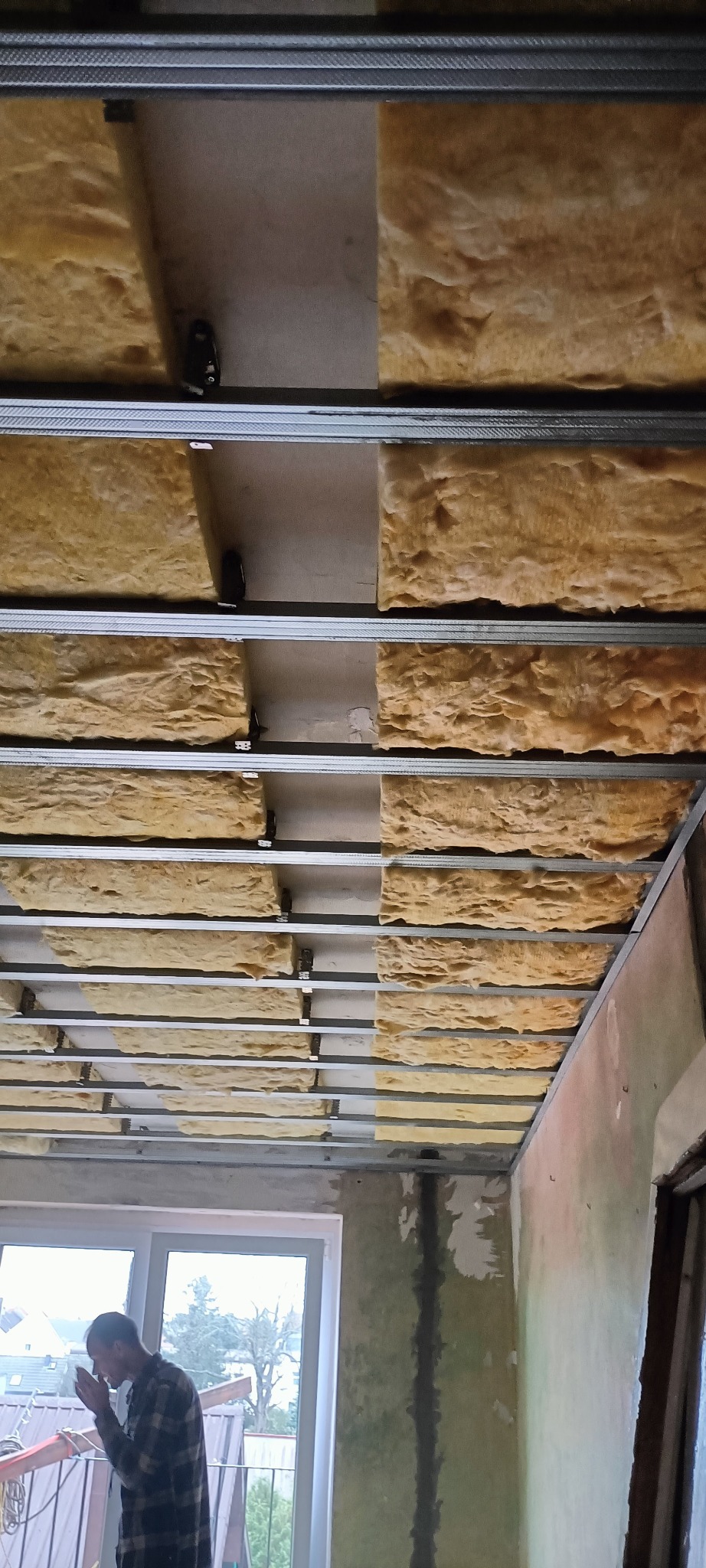 Montaż izolacji z wełny mineralnej na metalowym ruszcie sufitowym. Widoczna osoba w tle. Prace wykończeniowe w pomieszczeniu przed malowaniem.