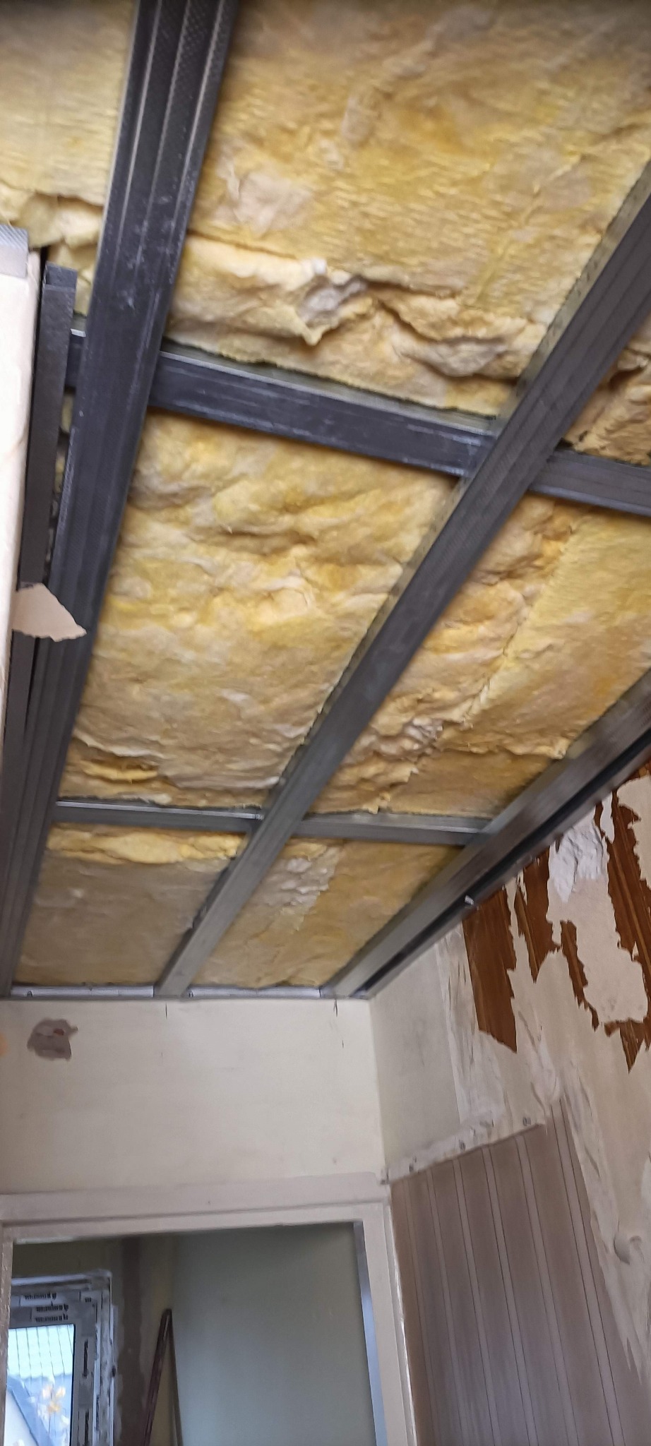 Montaż sufitu podwieszanego: metalowa konstrukcja z izolacją z wełny mineralnej, widoczne fragmenty starej ściany i ościeżnica drzwi. Etap prac wykończeniowych.