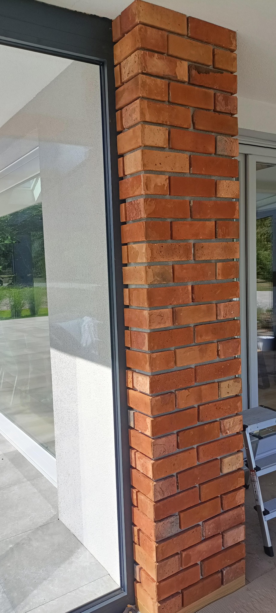 Wykończenie kolumny przy oknie cegłą klinkierową w odcieniach czerwieni i pomarańczy. Widoczny fragment drabiny. Elegancki detal wykończeniowy na zewnątrz budynku.