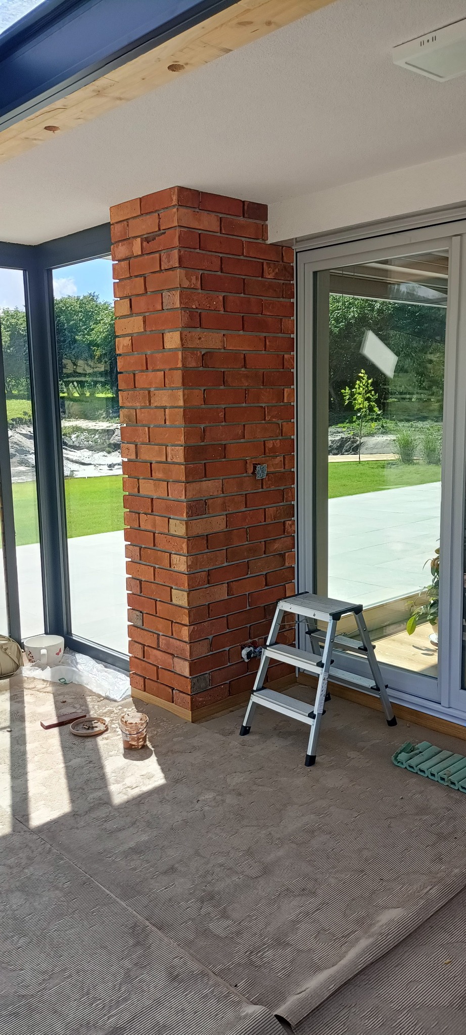 Murowany filar z czerwonej cegły w jasnym wnętrzu z dużymi oknami. Widoczna drabina, narzędzia i przygotowane podłoże. Częściowo wykończone wnętrze.