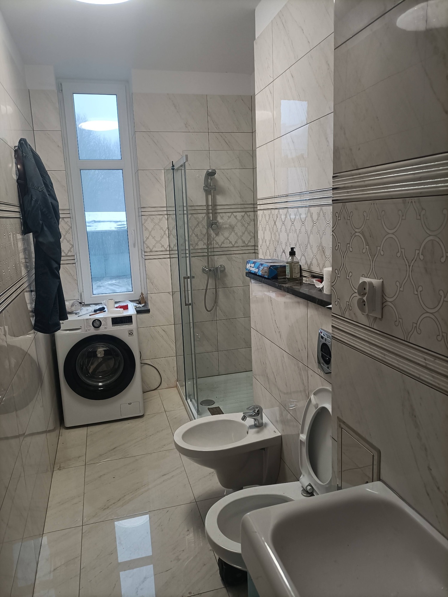 Jasna łazienka po remoncie z marmurowymi płytkami, kabiną prysznicową, pralką pod oknem i kompletem sanitarnym. Uwagę przykuwa wzór płytek na ścianach.