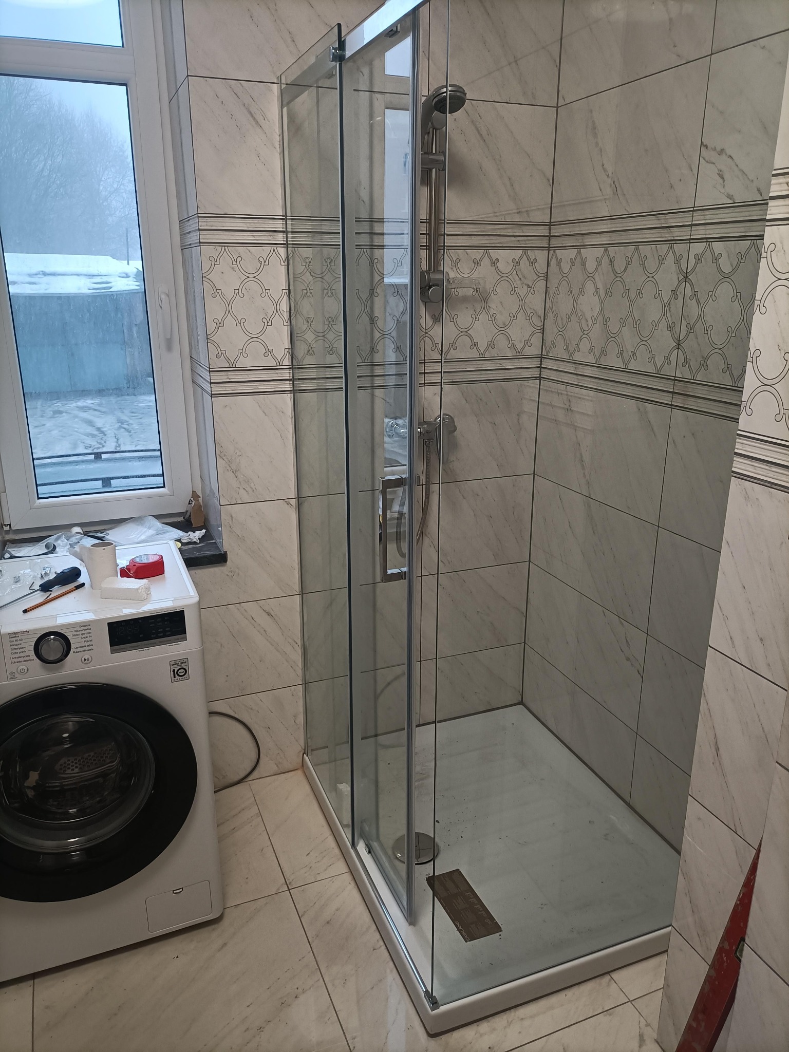 Wykończona kabina prysznicowa z marmurowymi płytkami i deszczownicą, widoczna pralka i okno z widokiem na zimowy krajobraz. Świeżo po remoncie.