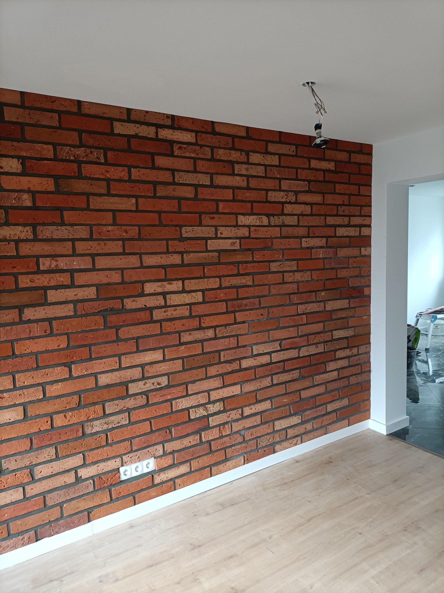 Ściana z czerwonej cegły w nowym wnętrzu, z widocznymi gniazdkami elektrycznymi i fragmentem lampy sufitowej. Podłoga wyłożona panelami.