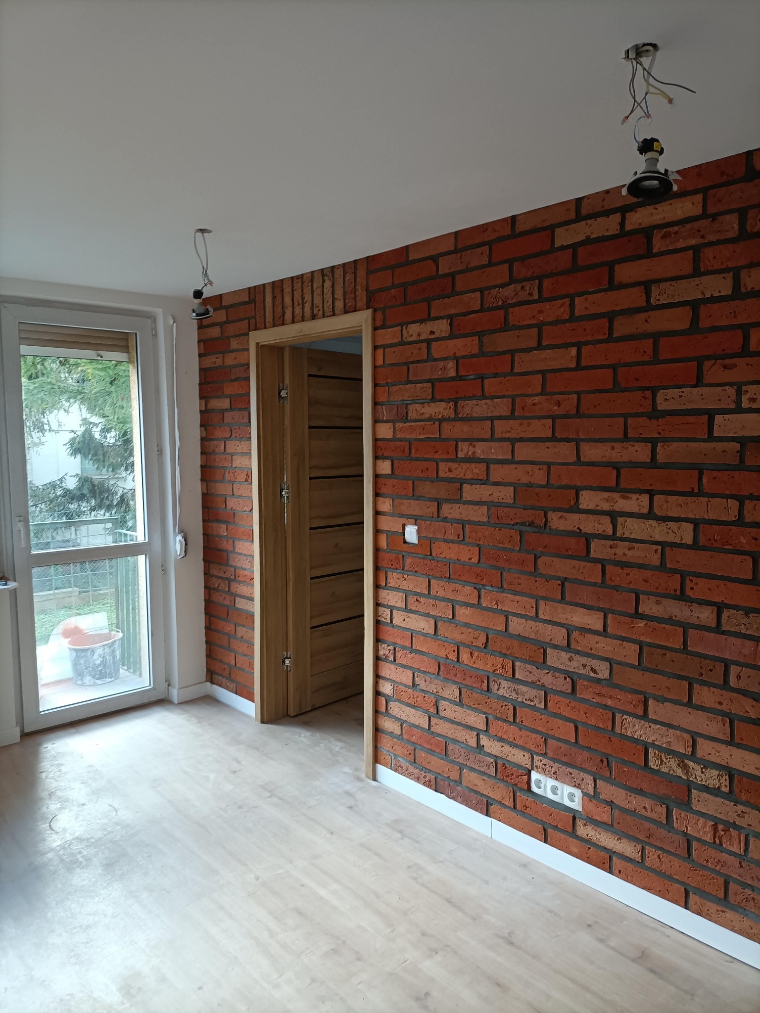 Pokój z ceglaną ścianą, drewnianą podłogą i ościeżnicą. Widoczne gniazdka elektryczne i niedokończone oświetlenie sufitowe. Okno z widokiem na zieleń.