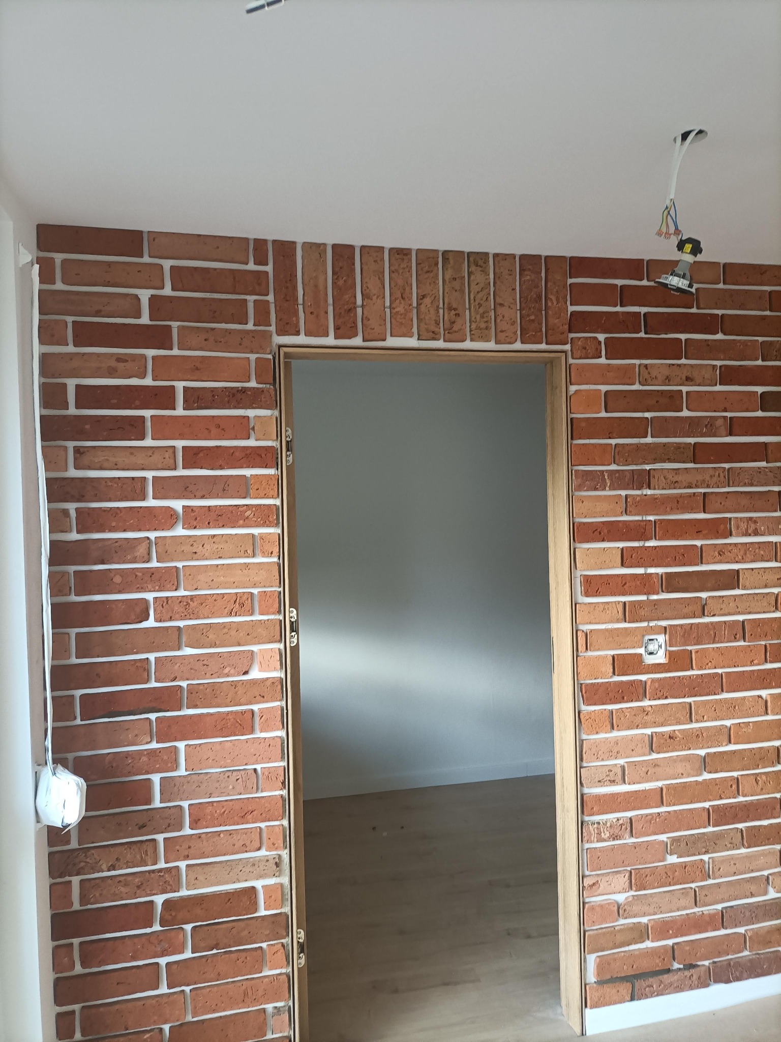 Ściana z cegły klinkierowej z drewnianą obudową drzwiową. Widoczna instalacja elektryczna i gniazdko. Jasne fugi, surowe wykończenie.