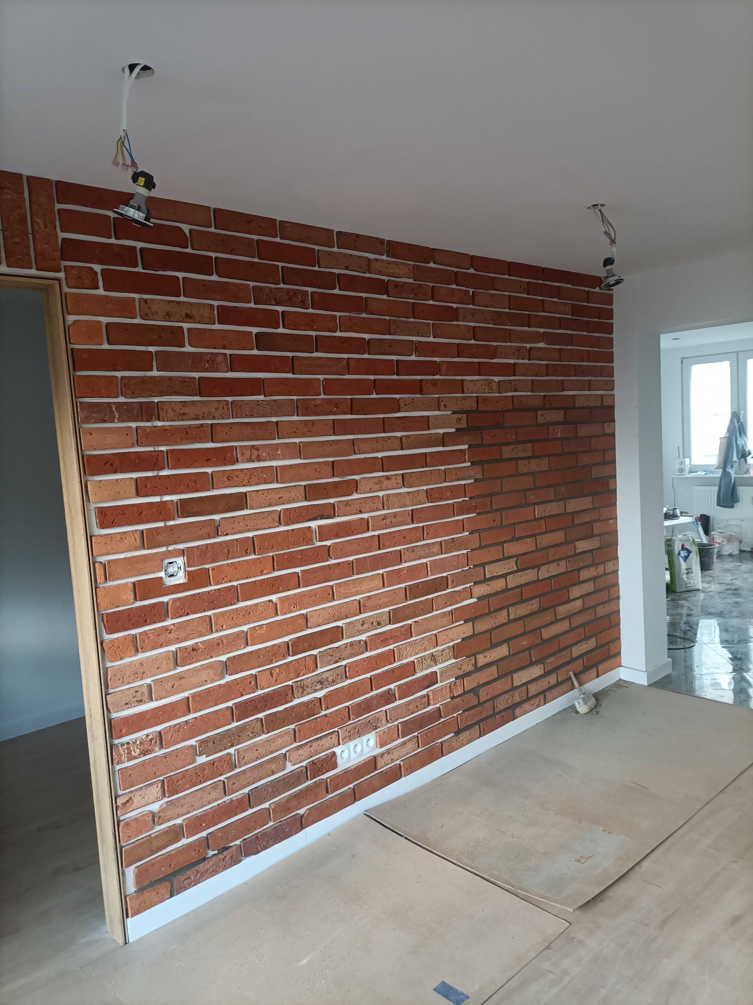 Ściana z cegieł klinkierowych w pomieszczeniu, widoczny fragment niezakończonej fugi, surowe przewody oświetleniowe, przygotowane podłoże.