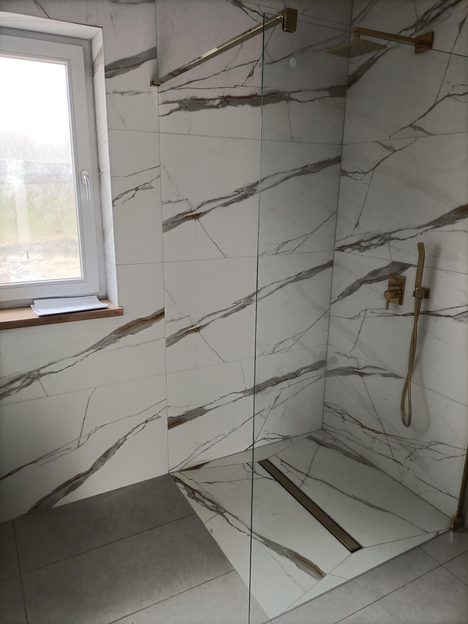 Wykończona kabina prysznicowa z odpływem liniowym, wyłożona płytkami imitującymi marmur z złotą armaturą. Widoczne okno z dokumentami na parapecie.