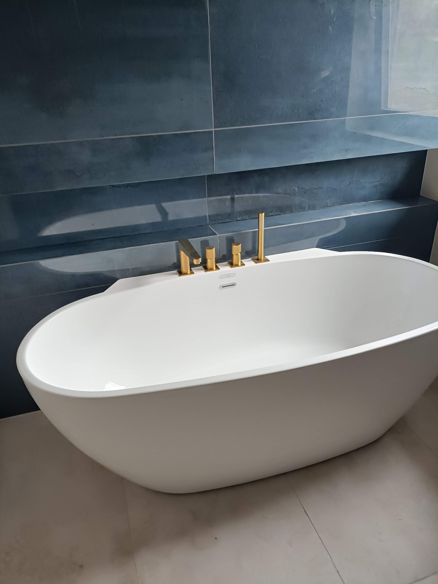 Wolnostojąca wanna Scandibath z złotą armaturą na tle ściany wyłożonej ciemnoniebieskimi płytkami w łazience po remoncie. Minimalistyczny design.