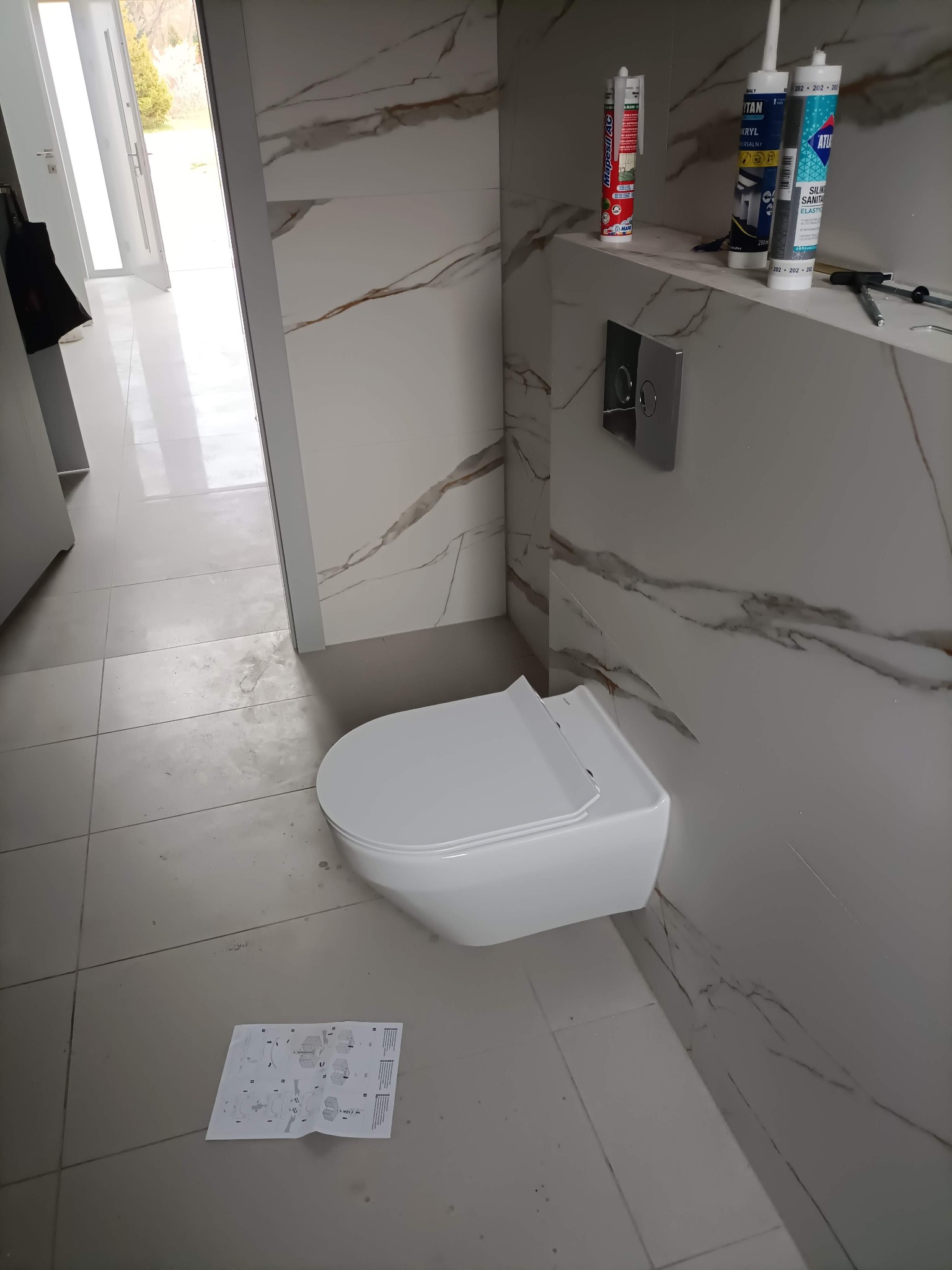 Montaż wiszącej toalety w łazience z marmurowymi płytkami. Widoczne narzędzia i instrukcja. Ukończony etap prac wykończeniowych.
