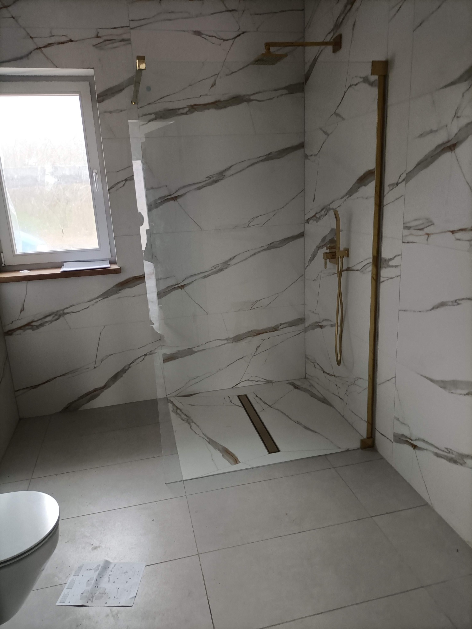 Wykończona łazienka z kabiną typu walk-in, białe płytki imitujące marmur z brązowymi żyłami, złota armatura i odpływ liniowy. Widoczne okno i część toalety.