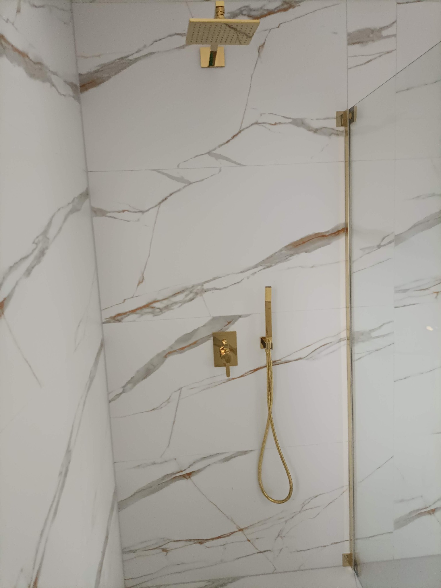 Kabina prysznicowa z białymi płytkami imitującymi marmur i złotą armaturą, w tym deszczownicą, baterią i słuchawką prysznicową. Szklana ściana kabiny z złotymi okuciami.