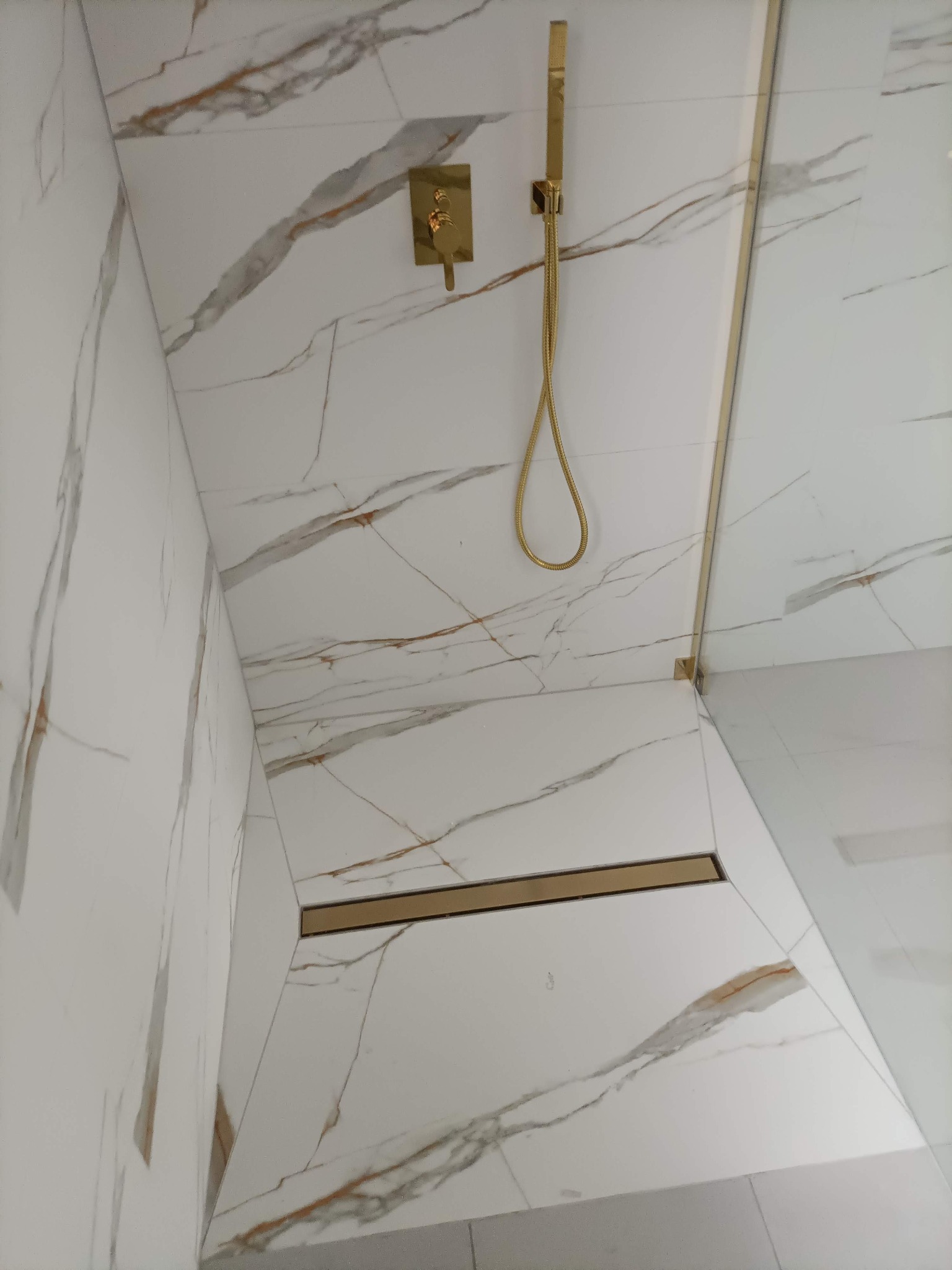 Białe płytki imitujące marmur z złotymi akcentami w kabinie prysznicowej, złota armatura i odpływ liniowy w Łebie. Nowoczesne wykończenie.