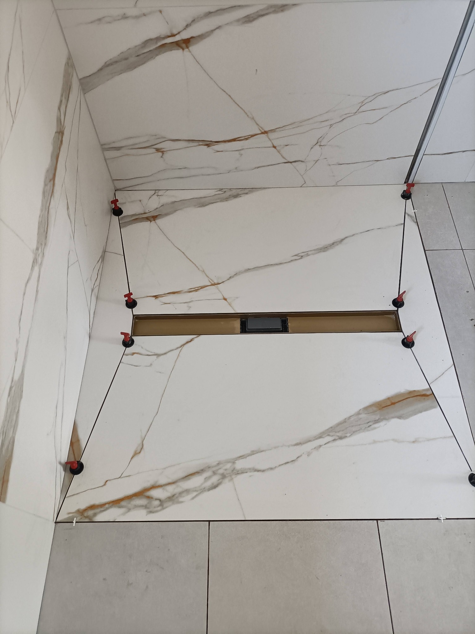 Białe płytki imitujące marmur z brązowymi żyłami, ułożone w kabinie prysznicowej. Widoczne systemy poziomowania płytek i odpływ liniowy.