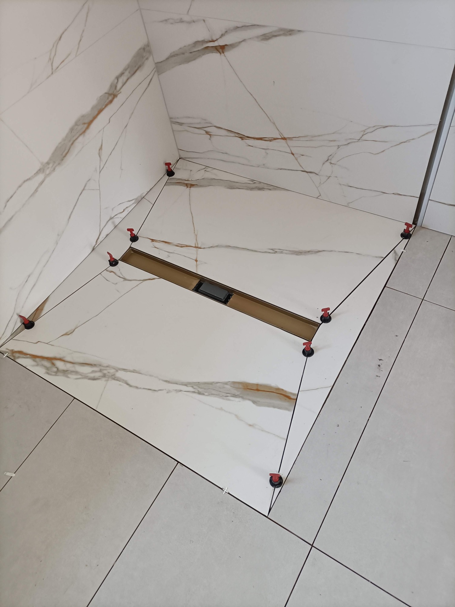 Ułożone płytki imitujące marmur w kabinie prysznicowej z odpływem liniowym. Widoczne klipsy poziomujące. Jasne płytki podłogowe.