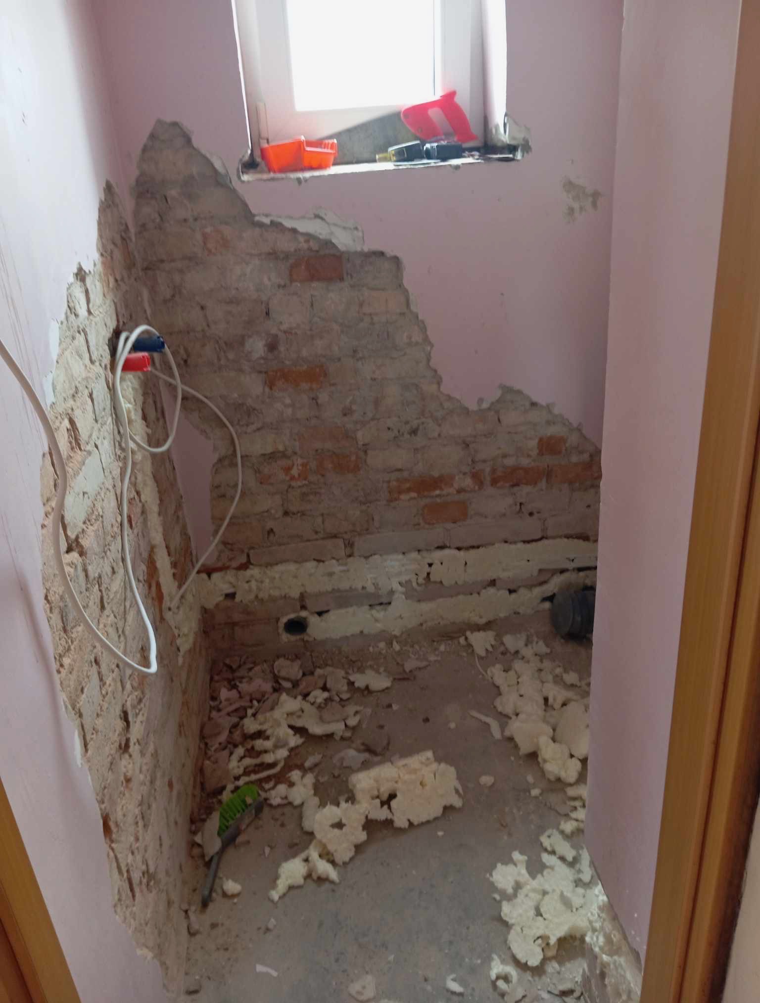 Odsłonięta cegła i instalacja w łazience w trakcie demontażu, widoczne resztki pianki montażowej na podłodze. Okno w tle z narzędziami.