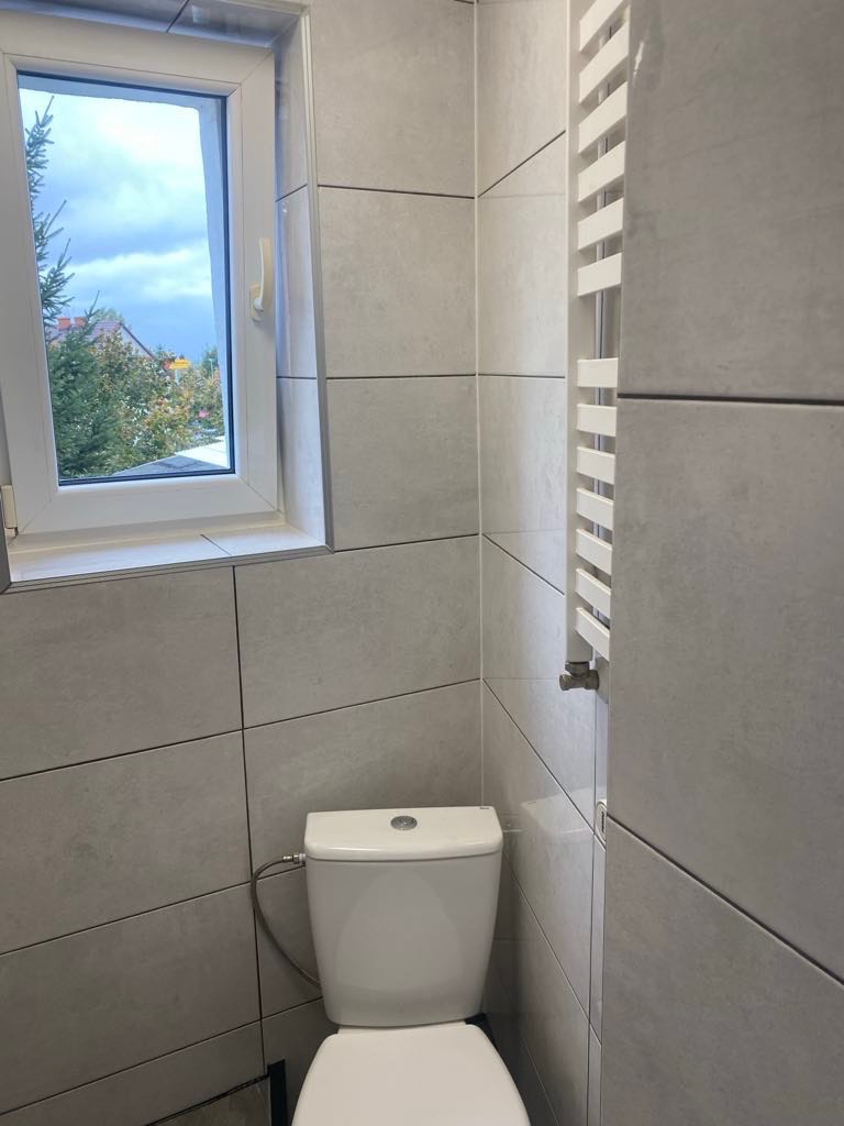 Jasna łazienka z toaletą, grzejnikiem drabinkowym i oknem z widokiem na zieleń. Płytki ceramiczne w odcieniach szarości ułożone regularnie na ścianach.