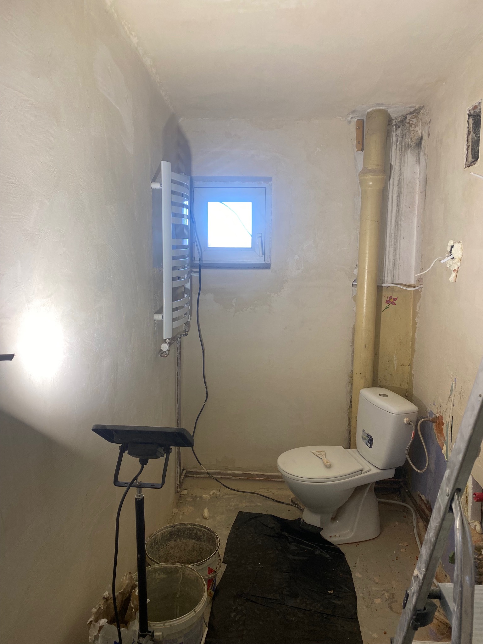 Łazienka w trakcie remontu: świeżo otynkowane ściany, zamontowany grzejnik, toaleta z narzędziem, widoczna instalacja i drabina malarska.