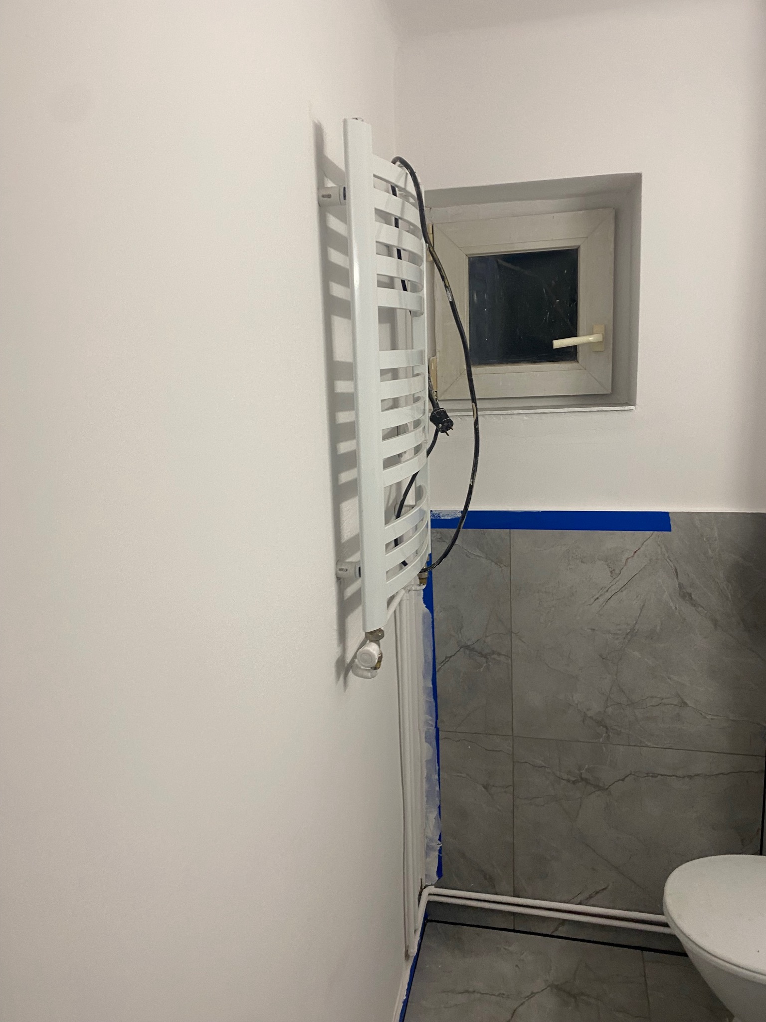 Biały grzejnik drabinkowy na ścianie łazienki z szarymi płytkami imitującymi marmur, zabezpieczonymi niebieską taśmą malarską. Widoczne okno i fragment toalety.