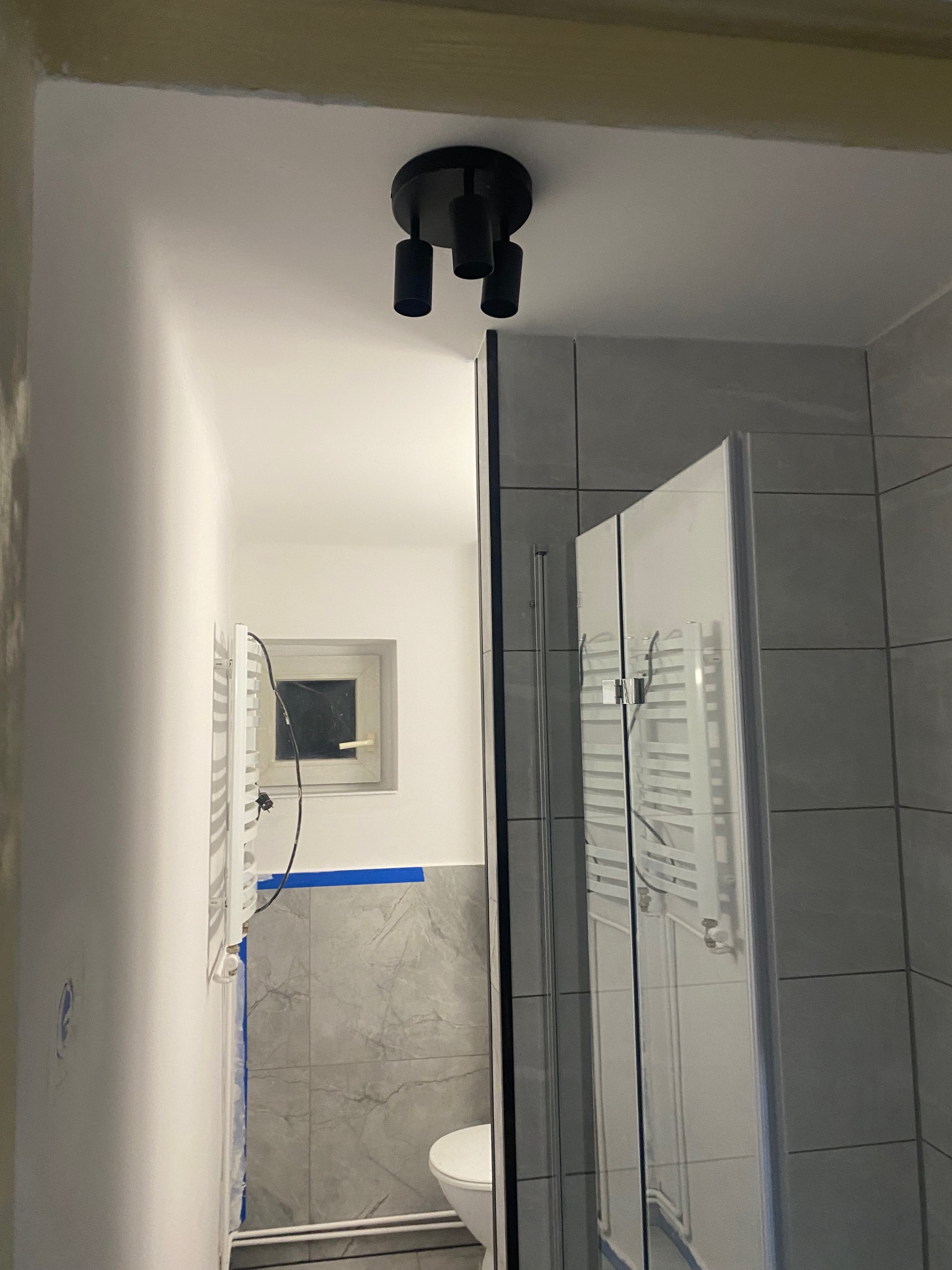 Nowoczesna łazienka po remoncie: kabina prysznicowa z szarymi płytkami, czarna lampa sufitowa i grzejnik na ścianie. Widok z perspektywy wejścia, minimalizm i funkcjonalność.