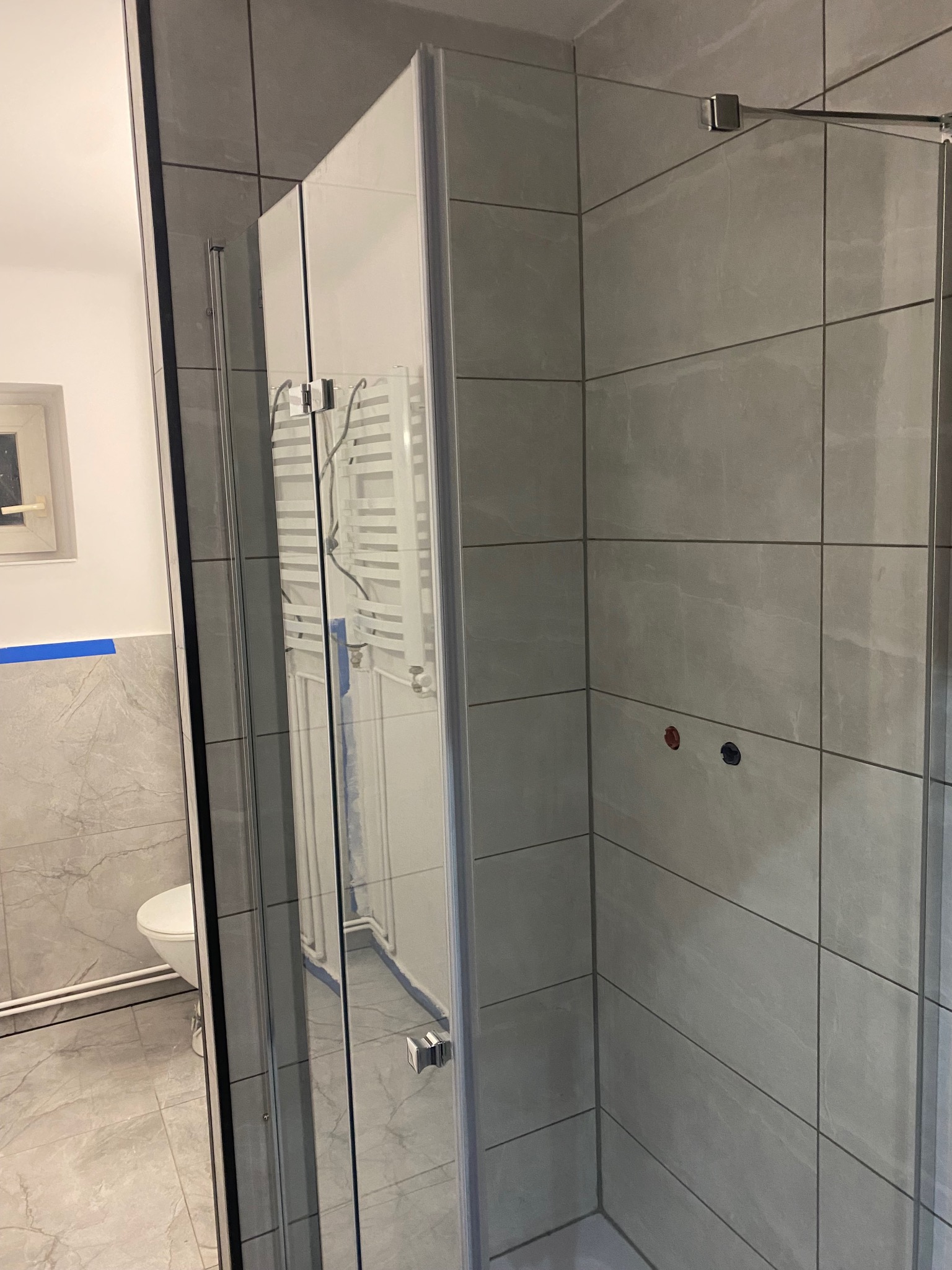 Szara, nowoczesna łazienka z kabiną prysznicową typu walk-in, widoczne płytki imitujące marmur i grzejnik drabinkowy. Minimalistyczny design.