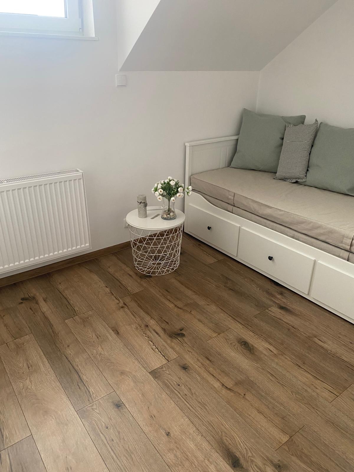 Jasna podłoga z paneli drewnopodobnych w pokoju na poddaszu. Biały grzejnik, stolik z kwiatami i kanapa z szufladami. Minimalistyczny wystrój wnętrza.