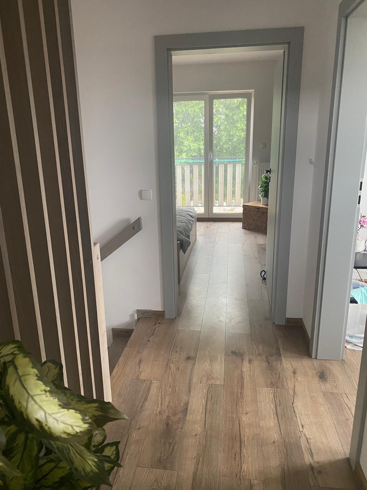 Parkiet ułożony w korytarzu w Strzałkowie, widok na balkon przez otwarte drzwi. Dekoracyjne lamele na ścianie i roślina w doniczce dodają wnętrzu charakteru.