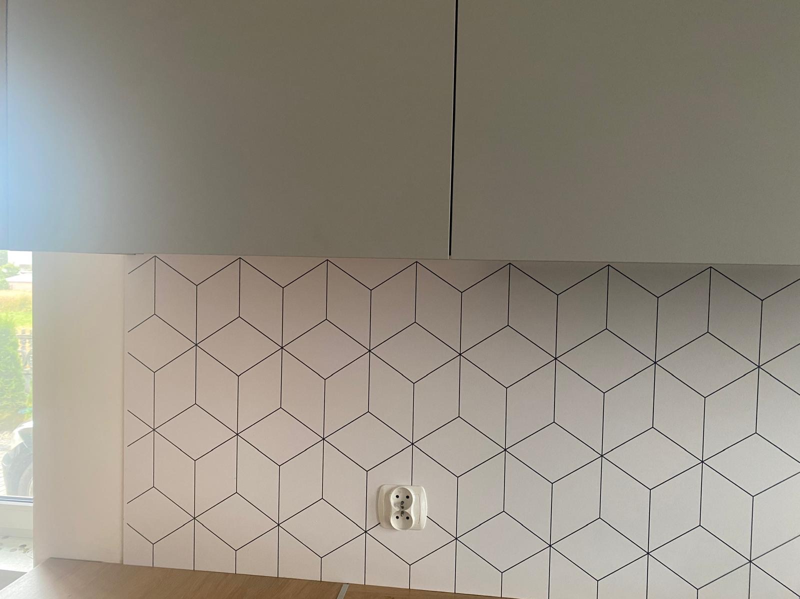 Białe płytki z geometrycznym wzorem 3D na ścianie nad blatem kuchennym, szare szafki wiszące i gniazdko elektryczne. Ujęcie z bliska, neutralne oświetlenie.