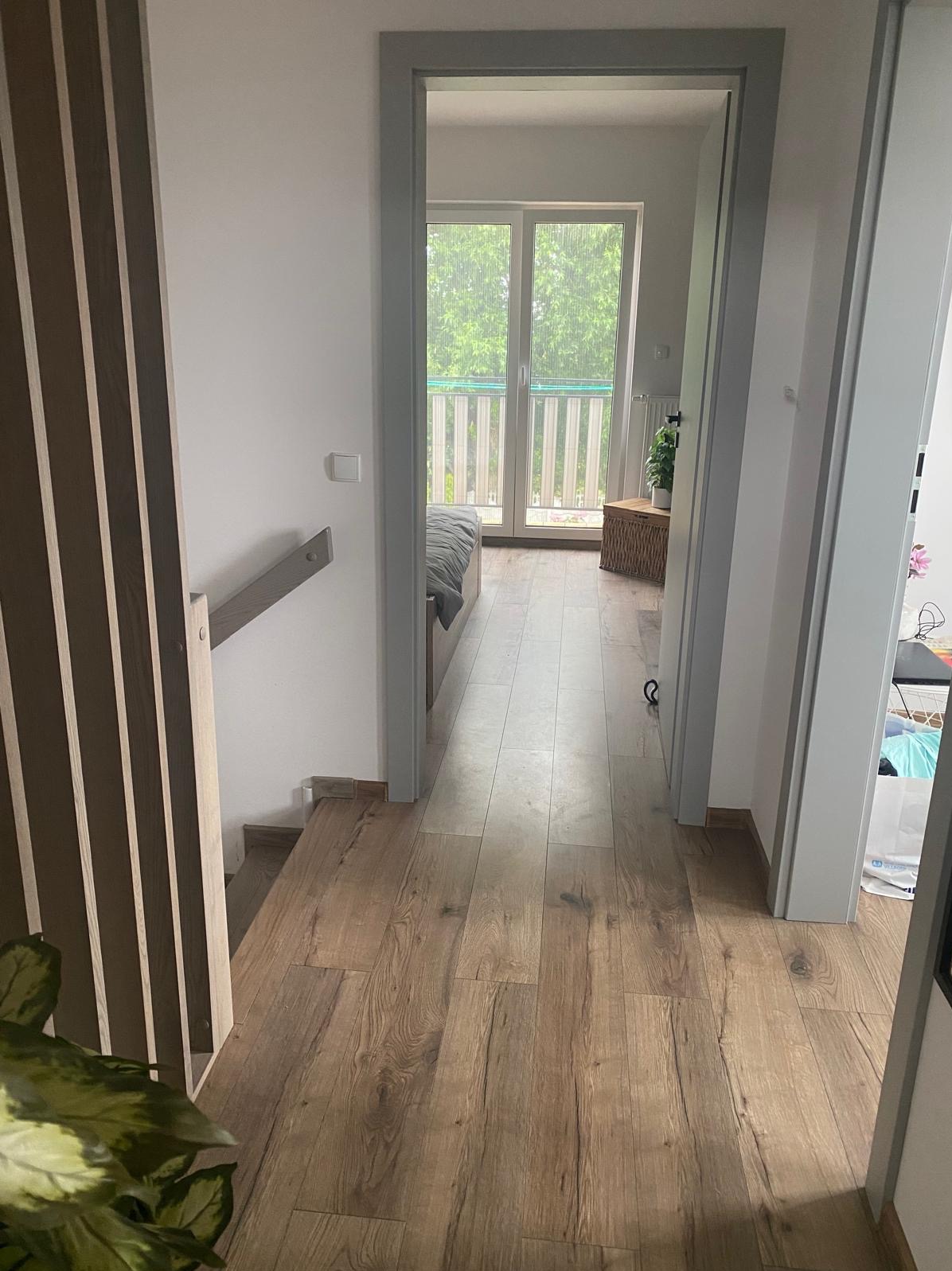 Jasne panele podłogowe prowadzą do pokoju z balkonem. Widoczne elementy wykończenia wnętrz: listwy przypodłogowe, drzwi z szarą ościeżnicą, fragment schodów.