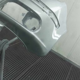 Rafał Ołdakowski - Pomalowany na srebrno, gładki zderzak samochodowy, umieszczony na metalowej kratownicy w lakierni. Widoczne mocowania i otwory na światła. Świeży lakier odbija światło.