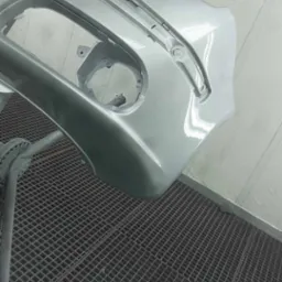 Pomalowany na srebrno, gładki zderzak samochodowy, umieszczony na metalowej kratownicy w lakierni. Widoczne mocowania i otwory na światła. Świeży lakier odbija światło.