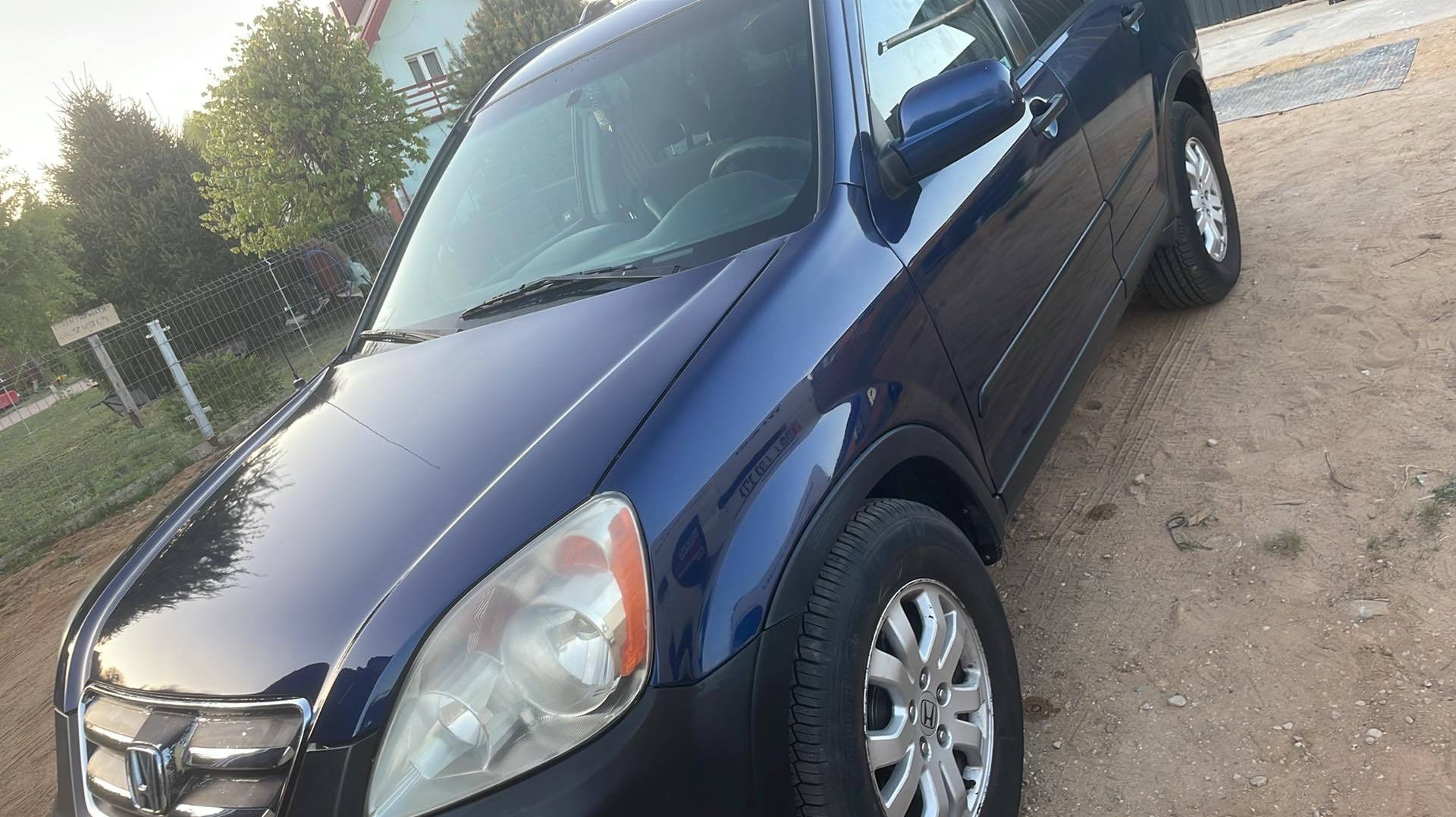 Błyszczący, granatowy SUV Honda CR-V na piaszczystym podłożu, z widocznymi felgami i fragmentem ogrodzenia w tle. Karoseria odbija światło słoneczne.