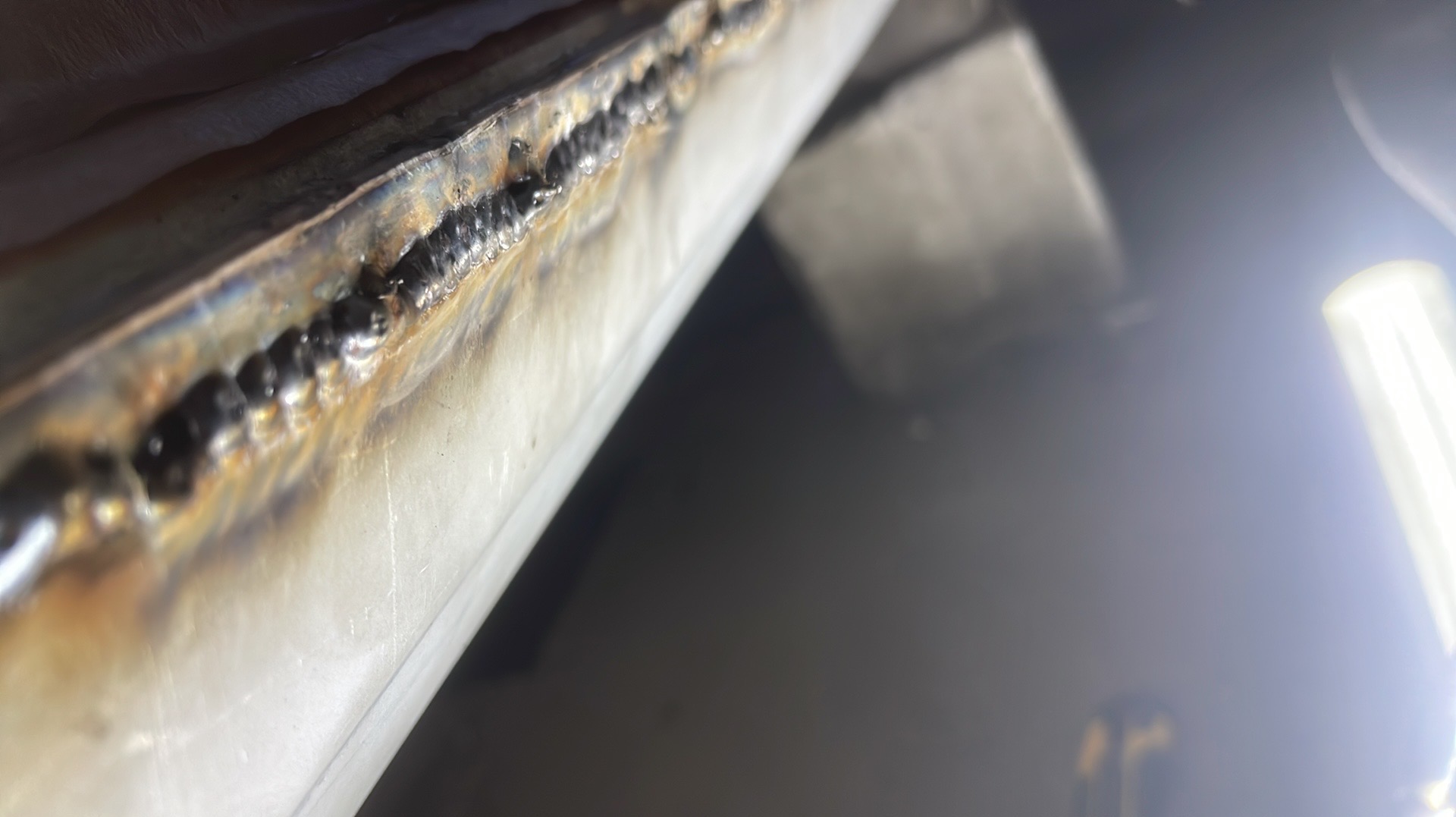 Detal spoiny na metalowej powierzchni, widoczne ślady ciepła i stopiony metal. Wyraźne, bliskie ujęcie spoiny, oświetlone jasnym światłem.