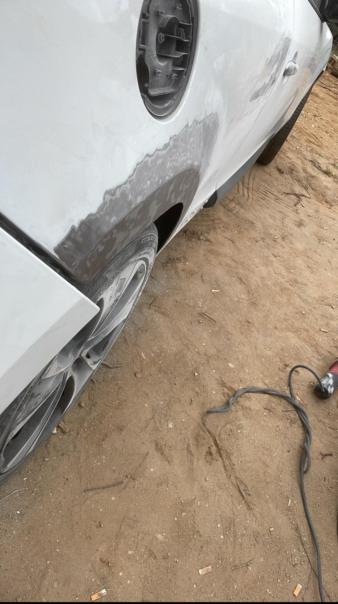 Biały samochód w trakcie naprawy blacharskiej: szpachlowana karoseria przy wlewie paliwa i kole. Widoczne narzędzia i ziemiste podłoże.