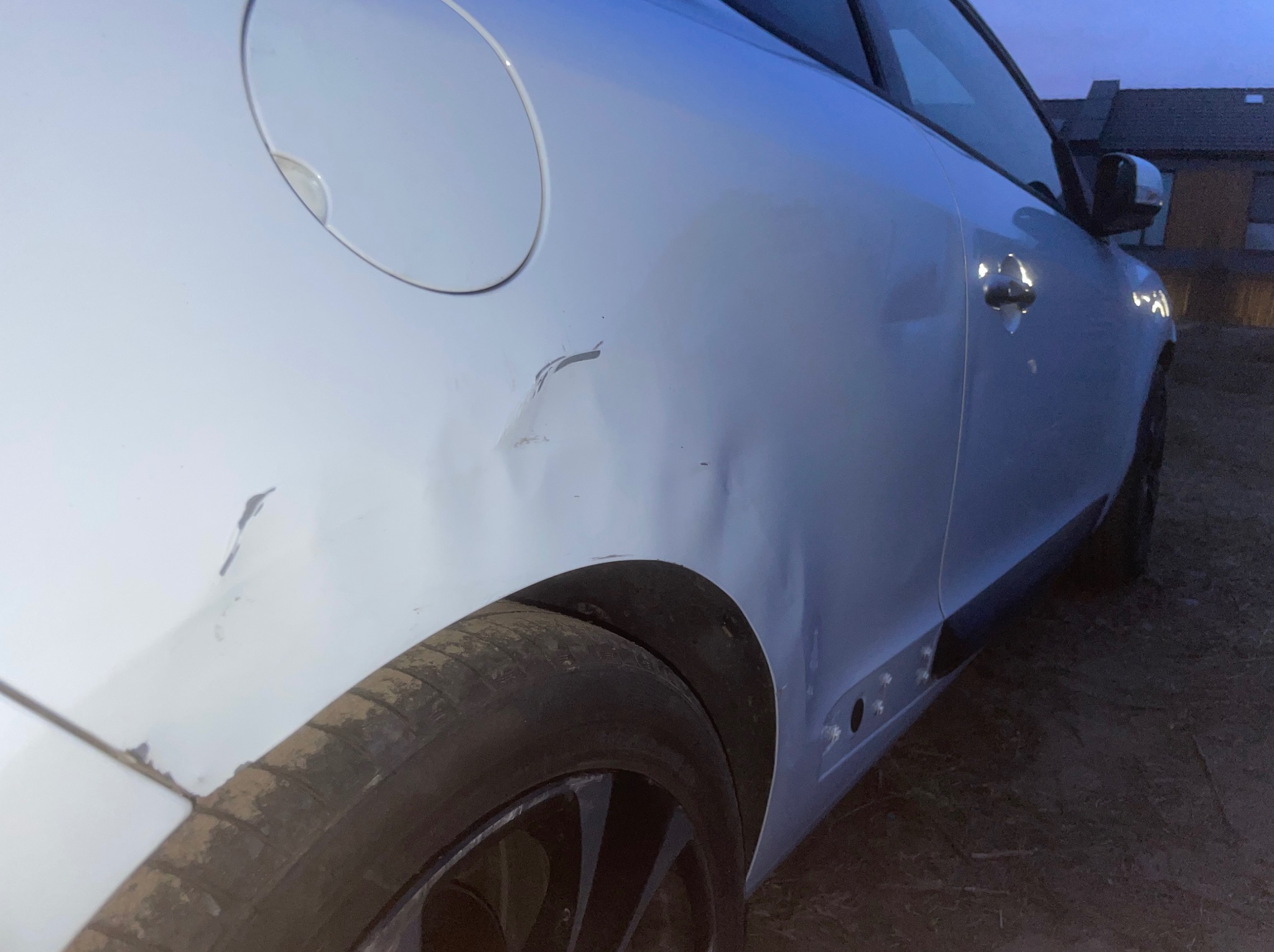 Uszkodzony bok srebrnego auta. Widoczne wgniecenia i zarysowania na karoserii w okolicy wlewu paliwa i tylnego nadkola. Brudna opona.