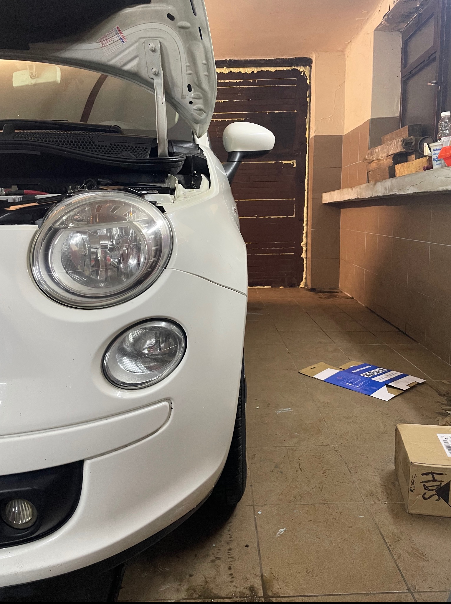 Biały samochód z otwartą maską w warsztacie. Widoczny reflektor, błotnik i fragment wnętrza silnika. Tło stanowią drzwi i półka z narzędziami.