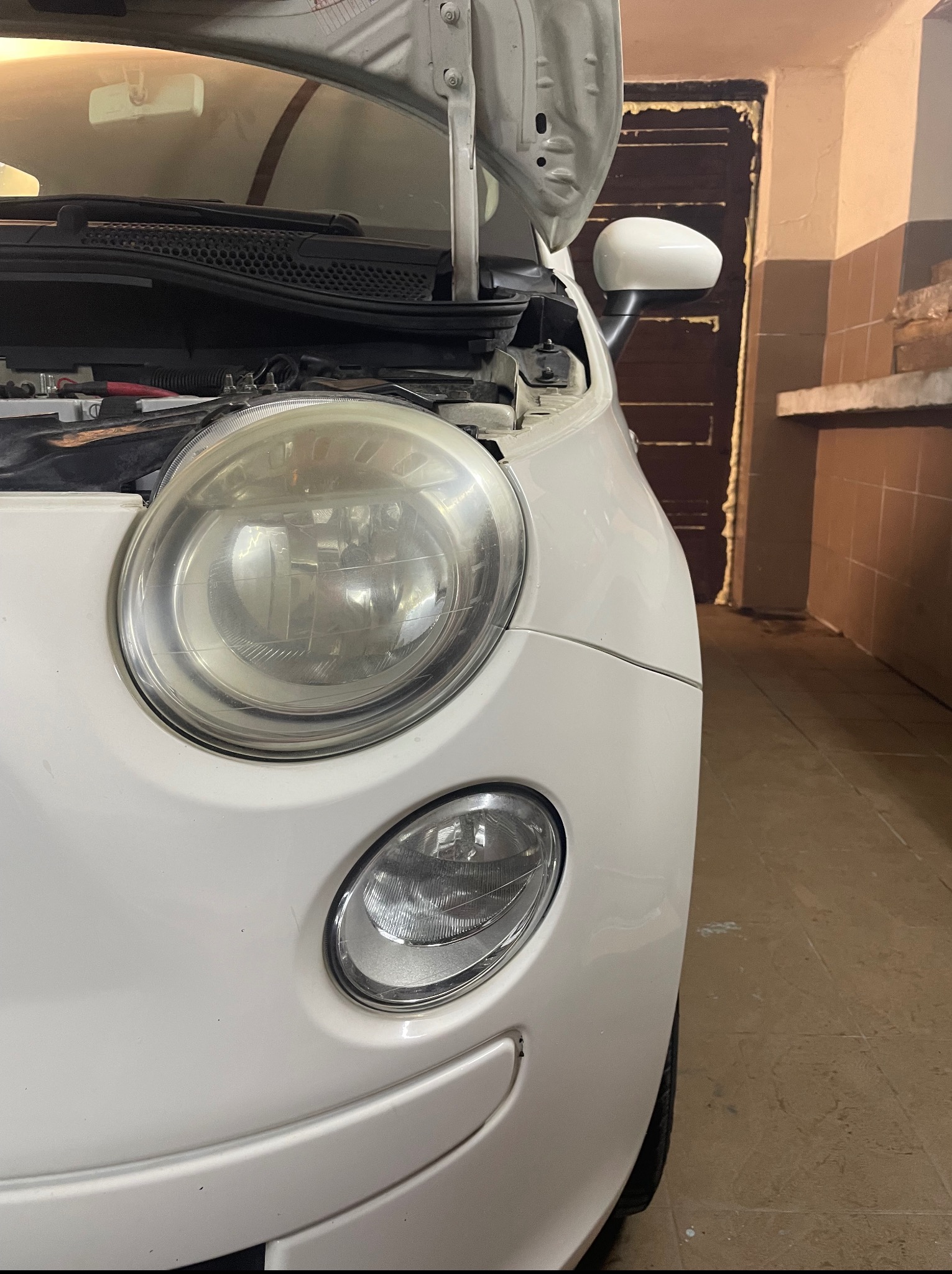 Biały Fiat 500 z otwartą maską, widoczny reflektor przedni, w tle ściana warsztatu. Auto przed naprawą/wymianą żarówki. Lekko przybrudzony klosz reflektora.