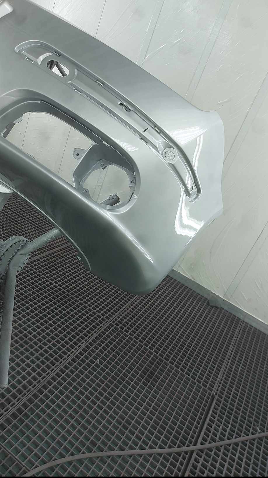 Pomalowany na srebrno, gładki zderzak samochodowy, umieszczony na metalowej kratownicy w lakierni. Widoczne mocowania i otwory na światła. Świeży lakier odbija światło.