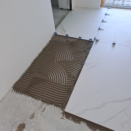 RENOPLAN Remonty i Wykończenia - Układanie płytek podłogowych z użyciem systemu poziomowania w Szczecinie. Widoczna nałożona zaprawa i płytki imitujące marmur, w tle drzwi i okno.