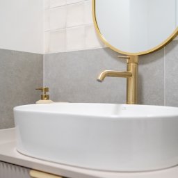 RENOPLAN Projekty i Wykończenia Wnętrz - Biała, owalna umywalka nablatowa z baterią w kolorze złotym, okrągłe lustro w złotej ramie, szare płytki na ścianie, minimalistyczny design łazienki.