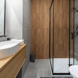 RENOPLAN Projekty i Wykończenia Wnętrz - Nowoczesna łazienka z drewnianą ścianą, szarą armaturą i kabiną prysznicową z czarnymi ramami. Umywalka nablatowa na drewnianej szafce, szare płytki podłogowe.