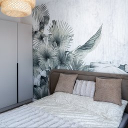 RENOPLAN Projekty i Wykończenia Wnętrz - Nowoczesna sypialnia z jasnoszarą szafą w zabudowie i łóżkiem z brązowym, zaokrąglonym zagłówkiem. Ściana z tapetą w szare liście palmowe, pleciona lampa sufitowa.