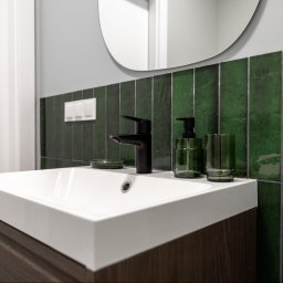 RENOPLAN Projekty i Wykończenia Wnętrz - Nowoczesna łazienka z białą umywalką, matową czarną baterią i szafką z ciemnego drewna. Ściana z błyszczących, pionowych, butelkowozielonych płytek z widoczną fugą, owalne lustro i zielone akcesoria.