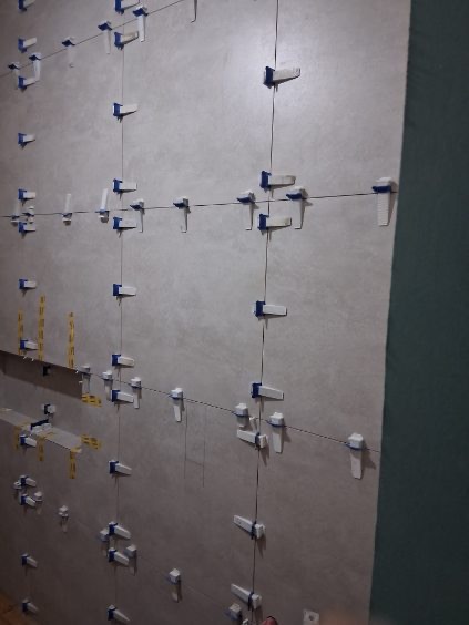 Ściana z szarych płytek, w trakcie montażu, z użyciem systemu poziomowania płytek z niebieskimi elementami. Widoczne linie sznurka wyznaczające poziom.