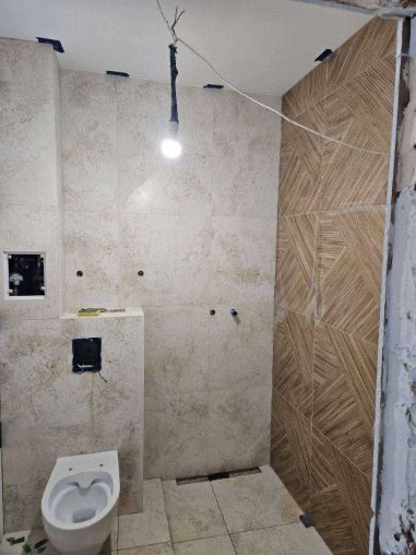 Wnętrze łazienki w trakcie montażu płytek, widoczny podwieszany sedes i fragment ściany z wzorem jodełki.  Oświetlenie tymczasowe.