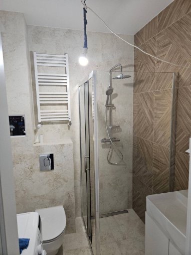 Nowoczesna łazienka z kabiną prysznicową i grzejnikiem drabinkowym. Uwagę przykuwa nietypowe oświetlenie w postaci wiszącej żarówki na kablu. Jasne płytki na ścianach.